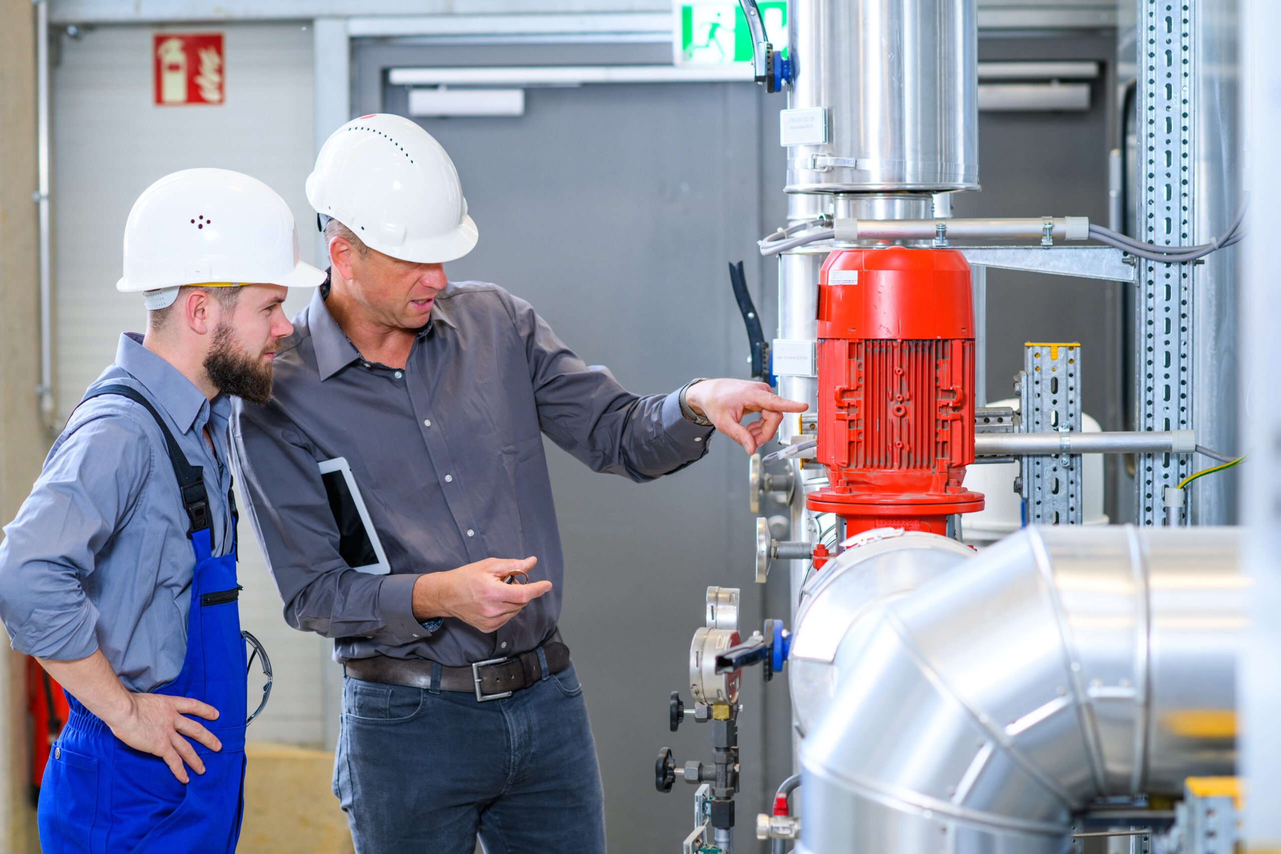 Two engineers, one pointing at a red motor, discuss maintenance procedures in a factory setting. Safety helmets are worn, indicating a working environment.