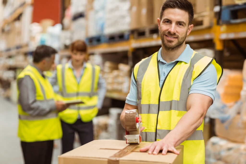 Warehouse worker in a safety vest smiling while taping a package, with colleagues discussing inventory in the background.