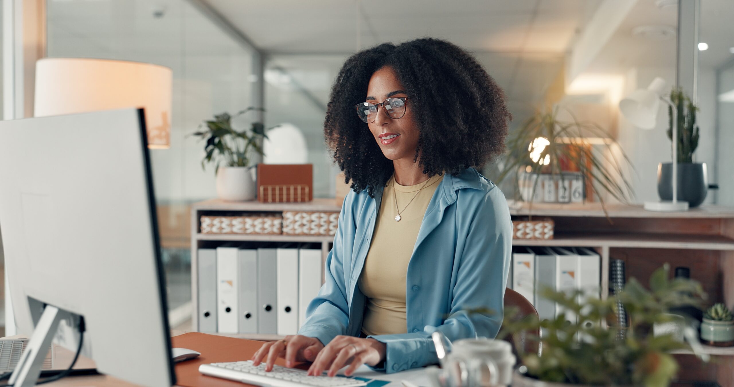 A woman with curly hair works at a desk, typing on a keyboard in a modern office. Plants and office supplies are visible in the background.