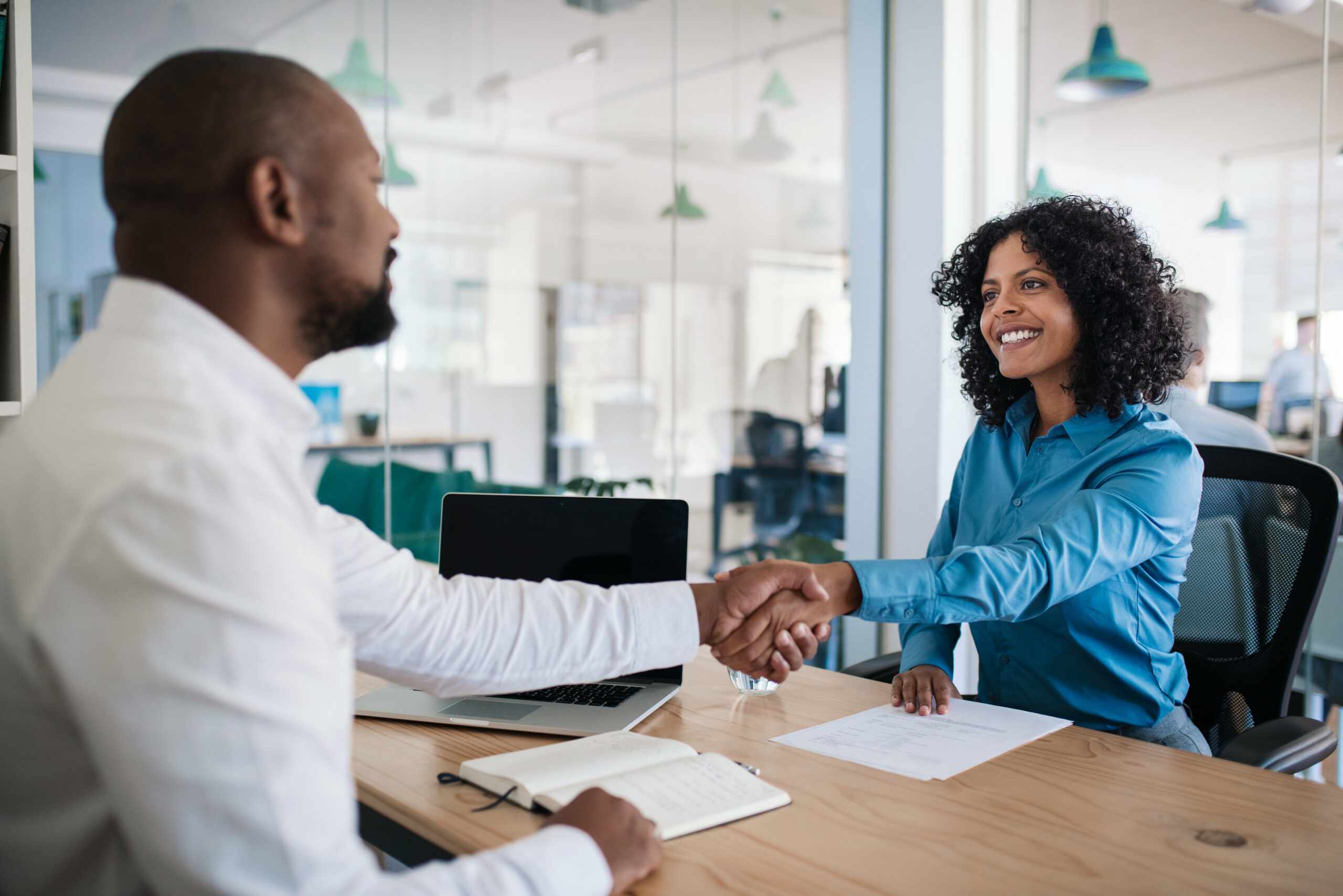 A woman with curly hair and a blue shirt shakes hands with a man in an interview setting, emphasizing a successful job interview moment.