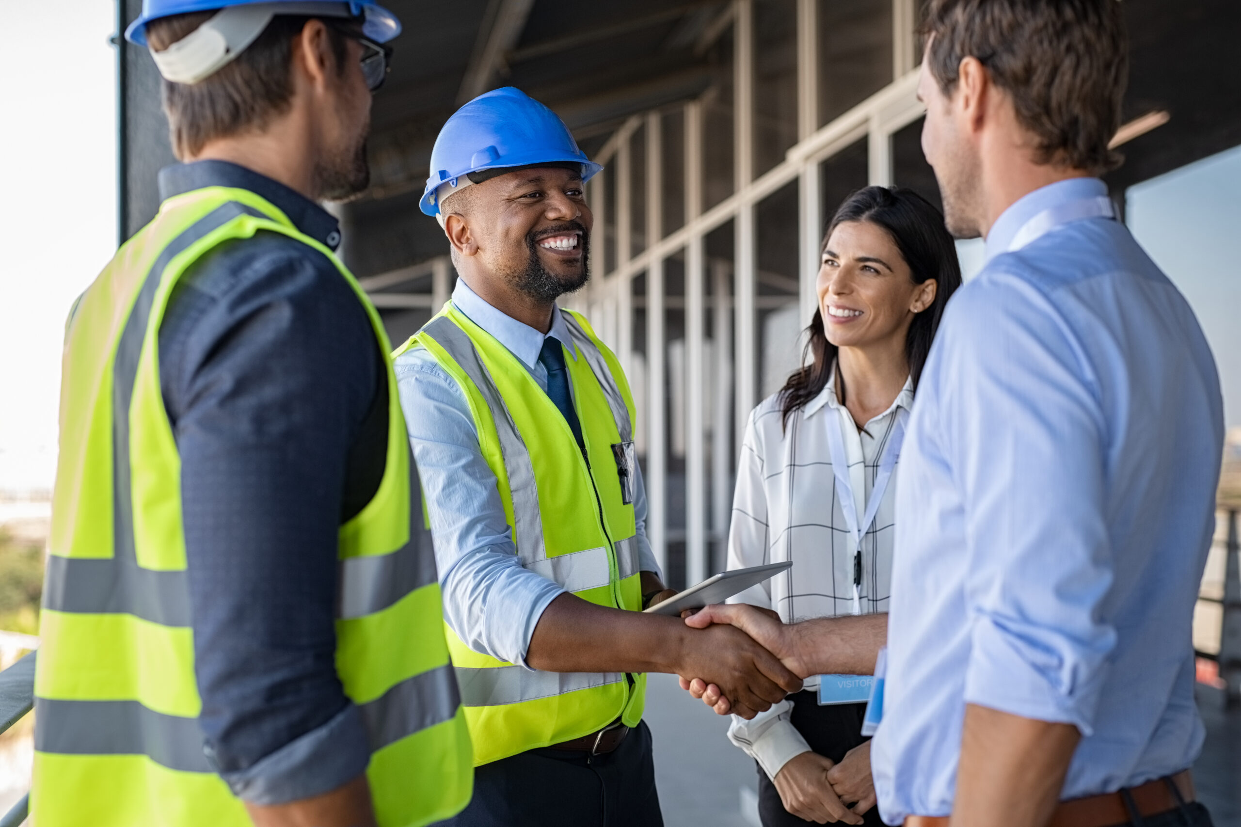 Construction professionals, including a man in a blue hard hat, shake hands and discuss project details in a bright, modern building setting.
