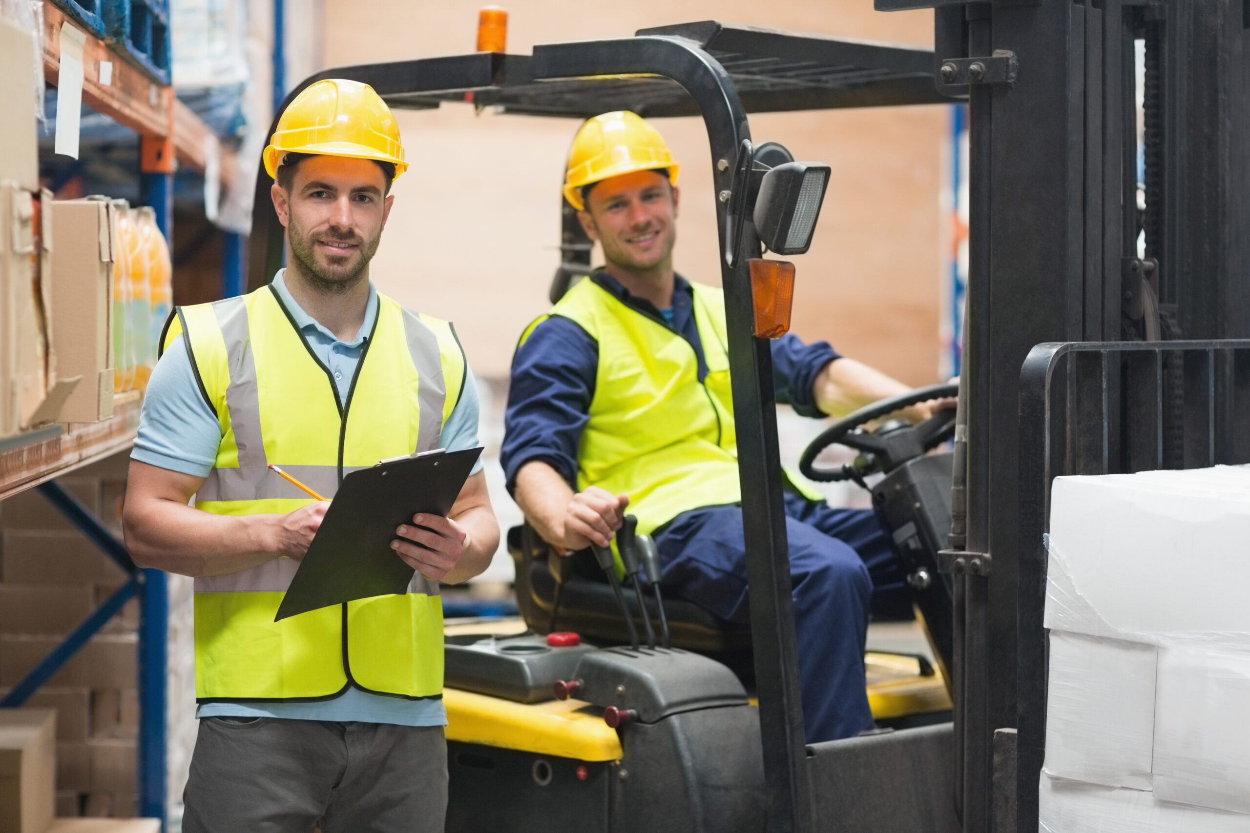 Warehouse workers in safety gear, one holding a clipboard and the other operating a forklift, oversee inventory management.